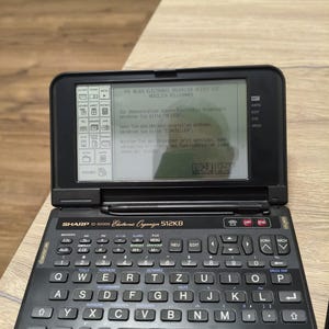 Può includere: Un organizer elettronico Sharp IQ-9200G nero con una capacità di memoria di 512KB. Il dispositivo è dotato di una tastiera QWERTY con etichette in tedesco, uno stilo riposto nella cerniera e uno schermo LCD monocromatico con un messaggio.