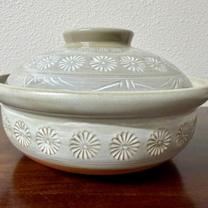 May include: A round ceramic lidded pot featuring a light beige glaze with a repeating white stamped daisy pattern around the base and the lid. The pot has two small side handles and a central knob on the lid, with a visible terracotta base.
