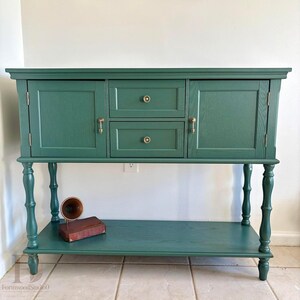 May include: A forest green wooden sideboard featuring two central drawers with round brass knobs, two side cabinets with brass drop pulls, and a lower open shelf. The piece is supported by four turned legs and includes a small vintage horn object.
