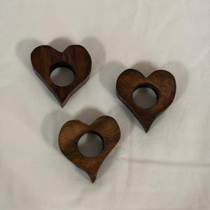 May include: Three dark brown wooden napkin rings, each shaped like a heart with a circular cutout in the centre. The wood grain is visible on the surface of each piece, showing a smooth, polished finish and a consistent, deep brown colour throughout.