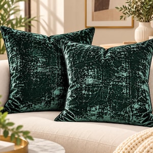 Può includere: Due cuscini quadrati di colore verde smeraldo intenso. Il tessuto presenta una consistenza in velluto stropicciato con un aspetto vissuto e maculato che crea variazioni di luce e ombra sulla superficie di ogni federa.