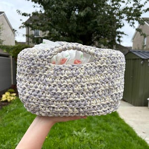 May include: A round, hand-crocheted basket crafted from thick, variegated yarn in shades of light grey, white, and cream. The basket features a sturdy, integrated handle opening at the top and contains several white items with small red accents.