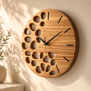 Könnte beinhalten: Eine runde Wanduhr aus Holz mit natürlicher Maserung. Die linke Seite weist zahlreiche unregelmäßige, organisch geformte Aussparungen auf, während die rechte Seite einfache schwarze lineare Stundenmarkierungen und drei dünne schwarze Zeiger zeigt.