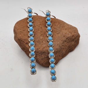 May include: A pair of long drop earrings featuring a vertical row of small, round, bright blue turquoise stones set in silver metal settings. Each earring is suspended from a simple silver hook, showcasing a linear design with twelve stones.