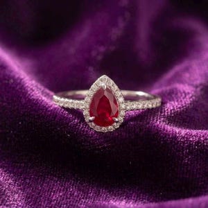 Pode incluir: Um anel com uma pedra central de rubi vermelho em forma de pera, rodeada por um halo de pequenos diamantes brancos. A banda é fina e metálica, também cravejada com pequenos diamantes brancos na metade superior, criando um aspeto brilhante.