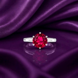 Può includere: Un anello in argento con una pietra preziosa rossa vibrante dal taglio rotondo a brillante, fissata con una montatura a sei griffe. La fascia è liscia e lucida, riflette la luce in modo nitido. L'anello è centrato, mostrando il colore rosso intenso.