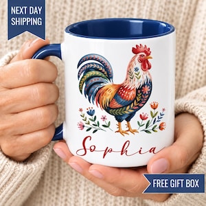 May include: A white ceramic mug with a dark blue interior and handle, featuring a colorful rooster illustration with floral accents and the name Sophia written in red cursive script below the bird on the front of the mug.