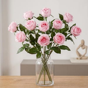 May include: A bouquet of eleven artificial light pink roses with layered petals and dark green leaves on long stems. The flowers are arranged in a clear glass vase with a cylindrical body and a narrow neck, showcasing the stems inside the glass.