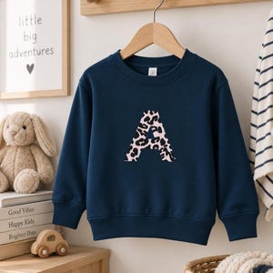 Könnte beinhalten: Ein langärmeliges, marineblaues Sweatshirt mit einem großen, mittig platzierten Buchstaben A. Der Buchstabe A ist mit einem rosa-schwarzen Leopardenmuster gefüllt. Das Kleidungsstück hat einen Rundhalsausschnitt sowie gerippte Bündchen.