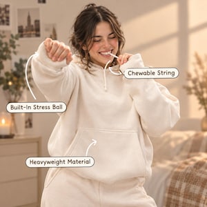 Könnte beinhalten: Ein cremefarbener Kapuzenpullover mit einem integrierten Stressball im Ärmel, einer kaubaren Schnur an der Kapuze und einer Kängurutasche vorne. Das Kleidungsstück besteht aus schwerem Material und ist mit Beschriftungen versehen, die diese Merkmale zeigen.