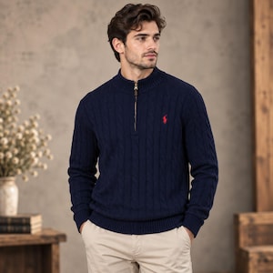 May include: A navy blue cable-knit sweater featuring a mock neck, a quarter-zip closure with a metallic zipper pull, and a small red embroidered logo on the left chest. The garment is paired with light beige trousers.