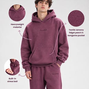 Può includere: Un set composto da felpa con cappuccio pesante e pantaloni della tuta in color prugna intenso. La felpa presenta una tasca a marsupio con una toppa sensoriale tattile, un logo ricamato sul petto e una pallina antistress nel polsino.