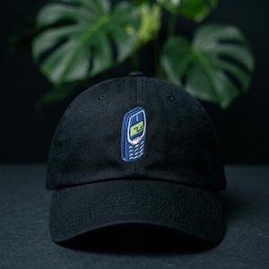 May include: A black baseball cap featuring a centred embroidered patch of a blue vintage mobile phone. The phone design includes a small screen with a light green display and a detailed keypad, positioned on the front panel of the cap.