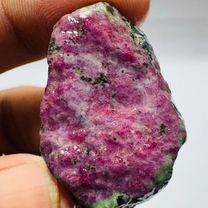 May include: A raw, unpolished gemstone featuring a vibrant magenta and pink crystalline surface with interspersed dark green and black mineral inclusions. The stone has an irregular, organic shape with a textured, multifaceted appearance.