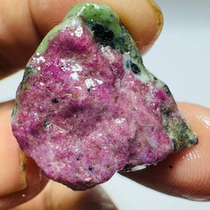 May include: A raw, unpolished mineral specimen featuring a vibrant magenta-pink centre surrounded by mottled green and black stone. The surface has a wet, glossy appearance with a rough, crystalline texture and irregular, jagged edges.