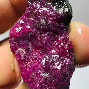May include: A raw, unpolished gemstone with a vibrant magenta and deep purple hue. The surface of the stone is textured, crystalline, and appears wet or glossy, showing natural mineral formations with dark, opaque inclusions at the top.
