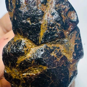 Può includere: Un esemplare minerale grezzo di colore marrone scuro e nero con una consistenza superficiale lucida e irregolare. La pietra presenta profonde fessure e crepe irregolari riempite con un deposito minerale terroso color giallo senape.