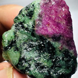May include: A raw, unpolished mineral specimen featuring a prominent section of vibrant magenta-pink crystal contrasted against a textured matrix of deep forest green and opaque black mineral inclusions with a naturally rough and uneven surface.