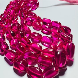 Puede incluir: Un hilo de cuentas translúcidas de color rosa intenso vibrante con un acabado suave y pulido. Las cuentas tienen forma ovalada y están ensartadas, con una borla de color rosa claro en el extremo del hilo, creando un componente de joyería brillante.