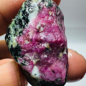 Op de afbeelding: Een ruwe, ongepolijste edelsteen met een levendig magenta-roze centrum omringd door donkergroene en zwarte mineraalinklusies. Het oppervlak van de steen heeft een kristallijn, getextureerd uiterlijk met zichtbare witte aders in het gesteente.