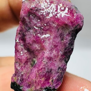 May include: A raw, unpolished ruby gemstone featuring a vibrant magenta and deep pink colour palette with natural crystalline textures and dark, opaque mineral inclusions throughout the irregular, elongated surface of the stone.