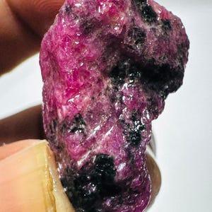May include: A raw, unpolished gemstone featuring a vibrant magenta and deep pink color palette with prominent, irregular black mineral inclusions throughout its textured, crystalline surface, showcasing a natural, jagged, and organic rock formation.