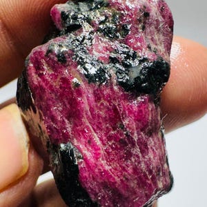 Pode incluir: Um espécime de pedra preciosa bruta não polida com uma estrutura cristalina magenta-rosa vibrante intercalada com inclusões minerais opacas de cor preta profunda. A superfície da pedra apresenta uma textura natural e rugosa com estrias visíveis.