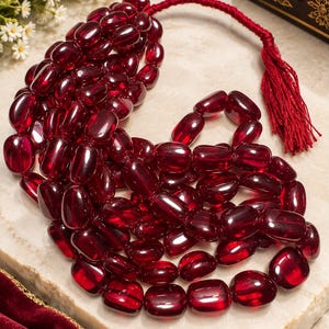 May include: A multi-strand necklace featuring numerous polished, deep red, translucent oval beads. The beads are strung on a dark red cord that terminates in a matching red tassel. The necklace is arranged in a dense, layered cluster.