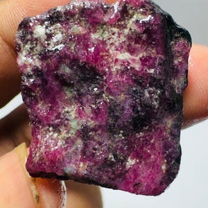 May include: A raw, unpolished gemstone with a deep magenta and dark purple color palette. The surface of the stone features a rough, crystalline texture with visible inclusions and varying levels of opacity throughout the irregular, jagged shape.