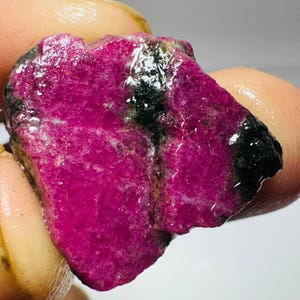 May include: A raw, unpolished gemstone with a vibrant magenta and deep pink hue, featuring a glossy, wet-looking surface texture. The stone displays irregular, jagged edges and prominent dark, black mineral inclusions running through the centre.