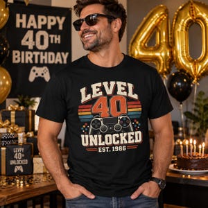 May include: A black short-sleeved t-shirt featuring a graphic print with the text "LEVEL 40 UNLOCKED EST. 1986" and a video game controller illustration. The design includes a retro-style sunset background in orange, yellow, and blue tones.