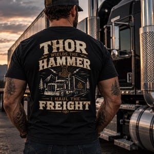 以下が含まれることがあります： 背面に「THOR WIELDS THE HAMMER I HAUL THE FREIGHT」の文字がプリントされた黒いTシャツ。デザインには、セミトレーラー、ハンマー、松の木、山々のイラストが、かすれたベージュ色で描かれています。