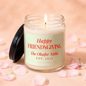 May include: A clear glass candle jar with a lit wick and a black lid. The label is light green with red text reading Happy FRIENDSGIVING The Okafor Table EST. 2021 and CANDLELETTERCO at the bottom. The candle is filled with white wax.