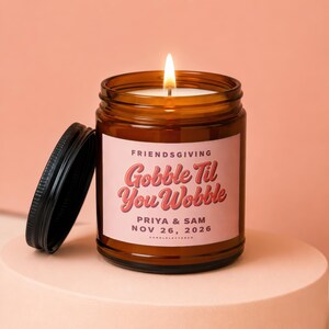 May include: A lit candle in an amber glass jar with a black lid. The label features the text "FRIENDSGIVING", "Gobble Til You Wobble", "PRIYA & SAM", and "NOV 26, 2026" in red and dark brown lettering on a light pink background.