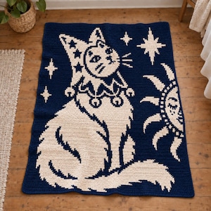 May include: A rectangular crochet blanket in dark navy blue and cream featuring a central cat wearing a jester hat with stars and a ruffled collar with bells. The design includes a sun with a face and several four-pointed stars in the background.
