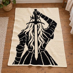 May include: A rectangular crochet blanket in cream with a large black graphic design of a figure wearing a crown and armor, holding a long sword upright with both hands. The design features a central sword blade and a detailed silhouette.