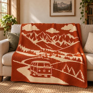 Puede incluir: Una manta de ganchillo rectangular con un diseño bicolor en naranja quemado y crema. El patrón muestra un paisaje con una furgoneta camper conduciendo por una carretera hacia picos montañosos, rodeada de pinos y nubes en el cielo.