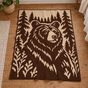 May include: A rectangular crochet blanket featuring a large, detailed bear head centered against a backdrop of pine trees. The design uses a two-tone color scheme of dark brown and cream yarn, creating a high-contrast graphic pattern.