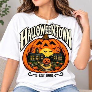 Peut inclure: Un t-shirt blanc avec un grand motif représentant une citrouille sculptée contenant une scène de ville avec des bâtiments, une petite citrouille d'Halloween et une lune jaune. Le texte HALLOWEENTOWN est arqué au-dessus et EST.1998 est imprimé en noir.