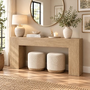 May include: A light wood console table with two cream-colored textured ottomans tucked underneath. On the table sits a beige lamp, a stack of books titled Kinfolk and At Home, a small bowl, a reed diffuser, and a vase with green leafy branches.
