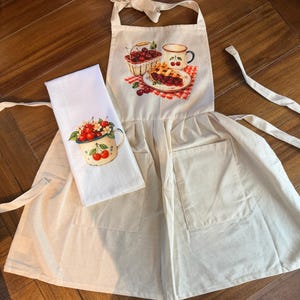 May include: A cream-colored apron with two front pockets and a matching white kitchen towel. The apron features a graphic of a basket of cherries, a cherry-patterned mug, and a cherry pie. The towel displays a mug filled with red cherries.