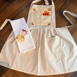 May include: A cream-colored apron featuring a printed Cherry Pie recipe with illustrations of cherries and a slice of pie, paired with a white kitchen towel displaying a graphic of nested measuring cups tied with a red ribbon.