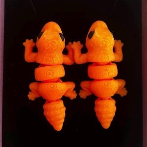 May include: Two bright orange articulated lizard figurines with visible 3D printed layer lines. Each lizard features a textured body, four small legs, and a segmented tail. The figures have dark, round eyes and are positioned side by side.
