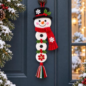 May include: A crocheted snowman wall hanging featuring a black top hat with a white snowflake, a bright orange carrot nose, pink cheeks, and a red scarf. The body consists of three white circular segments with black buttons, green holly, and a tassel.