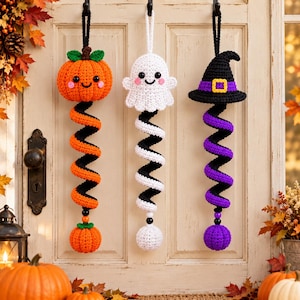 May include: Three hanging crochet decorations featuring a pumpkin, a ghost, and a witch hat, each attached to a long, colourful spiral tail ending in a small matching sphere. The pumpkin is orange, the ghost is white, and the hat is black and purple.