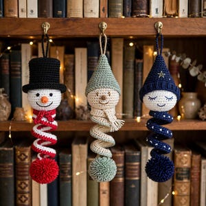 May include: Three crocheted ornaments hanging from hooks. The left ornament is a snowman with a black top hat, orange nose, and red and white spiral body. The center is a figure with a sage green hat and spiral body. The right is a figure with a navy blue hat and star-patterned spiral body.