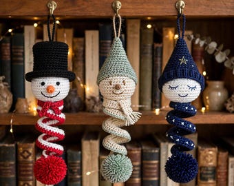 May include: Three crocheted ornaments hanging from hooks. The left ornament is a snowman with a black top hat, orange nose, and red and white spiral body. The center is a figure with a sage green hat and spiral body. The right is a figure with a navy blue hat and star-patterned spiral body.