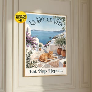 Peut inclure: Une affiche murale encadrée représentant une scène côtière méditerranéenne avec des bâtiments blancs, des fleurs violettes et des oliviers. Un chat tigré orange se repose sur un chemin en pierre. Le texte indique LA DOLCE VITA et Eat. Nap. Repeat.