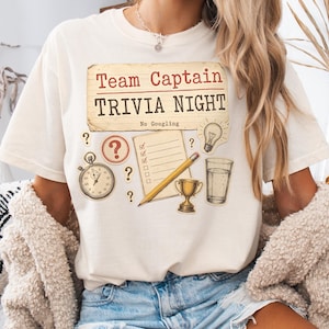Puede incluir: Una camiseta de color crema con un gráfico que incluye el texto Team Captain TRIVIA NIGHT y No Googling. El diseño muestra un cronómetro, un signo de interrogación, una lista con un lápiz, una bombilla, un trofeo y un vaso de agua.