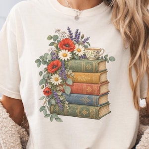 May include: A cream-colored t-shirt featuring a graphic print of a stack of five vintage-style books in shades of green, yellow, red, and blue, topped with a small floral teacup and a vibrant arrangement of red poppies and daisies.
